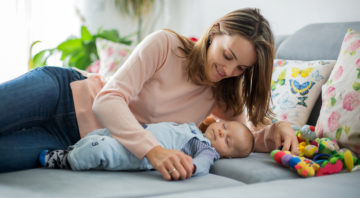 Cute baby boy and his mother, lying on the couch in living room, playing with toys, activity for early infant development