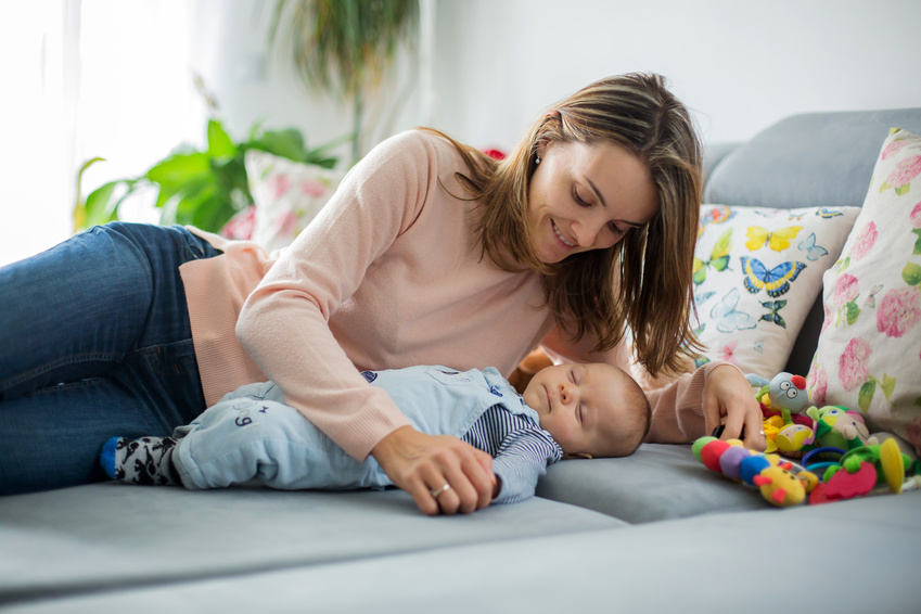 Cute baby boy and his mother, lying on the couch in living room, playing with toys, activity for early infant development